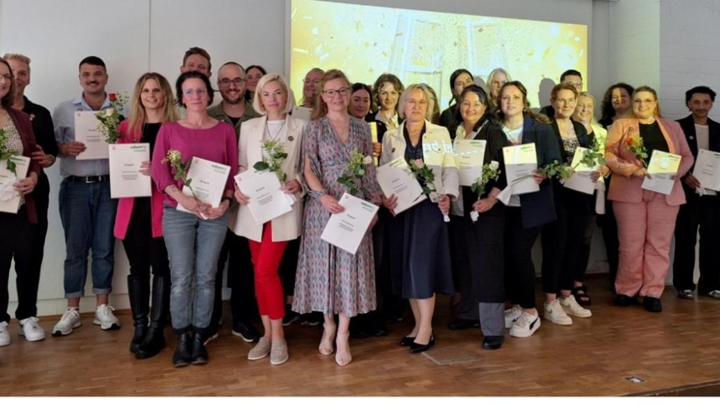 Absolvent:innen stehen strahlend auf dem Podium der Aula und präsentieren ihre Zertifikate