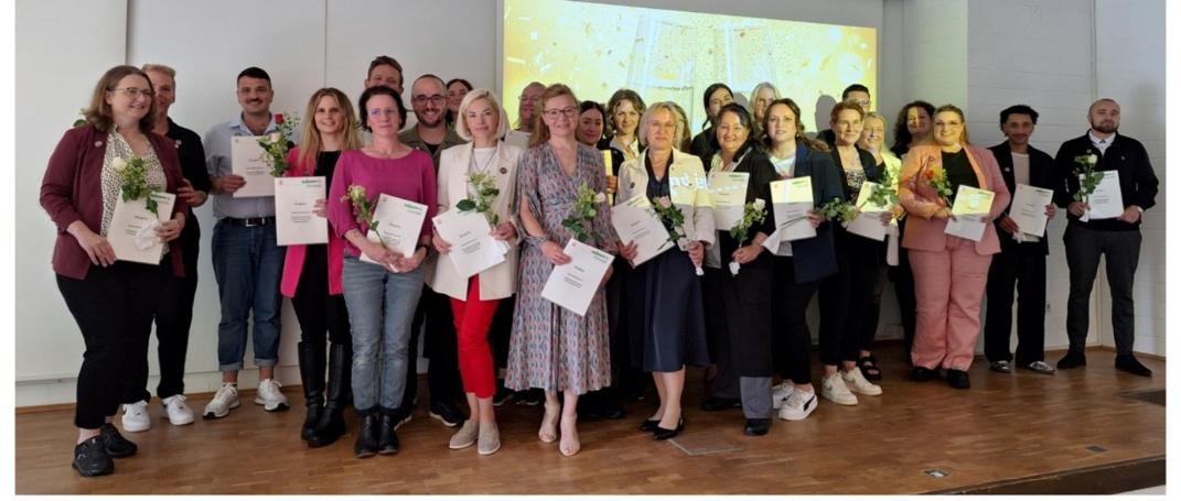 Absolvent:innen stehen strahlend auf dem Podium der Aula und präsentieren ihre Zertifikate