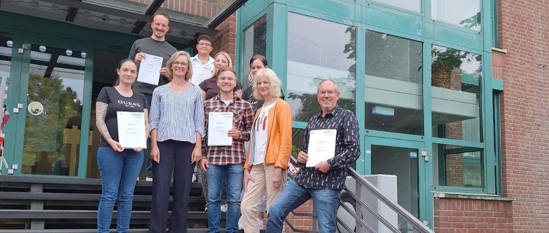 Pflegeprozessmanger:innen im Herbst 2026 Acht glückliche Absolvent:innen stehen mit der Kursleiterin Frau Tubbesing gemeinsam vor der Caritas-Akademie Köln-Hohenlind