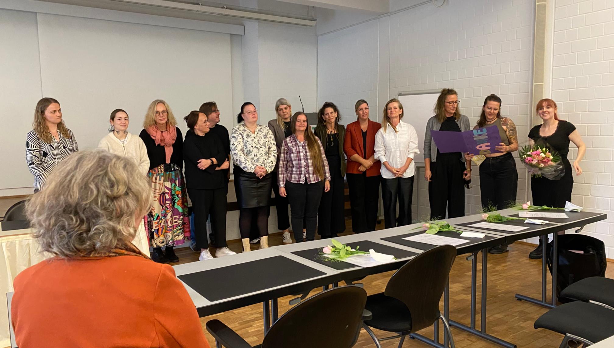 15 Teilnehmende stehen auf der Bühne und eine Dame sitzt im Auditorium davor.